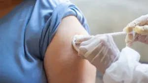 Medicinos darbuotojas su pirštinėmis leidžia vakciną į moters petį, naudodamas vienkartinį švirkštą.