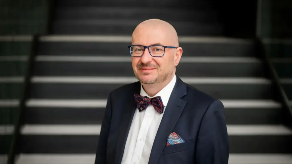 Plikai skustas vyras su akiniais, tamsiu kostiumu ir marga peteliške stovi laiptų fone.