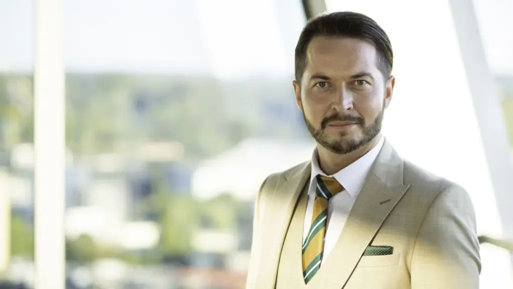 Vidutinio amžiaus barzdotas vyras šviesiu kostiumu stovi šalia didelio lango šviesioje patalpoje.