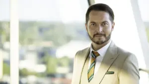 Vidutinio amžiaus barzdotas vyras šviesiu kostiumu stovi šalia didelio lango šviesioje patalpoje.