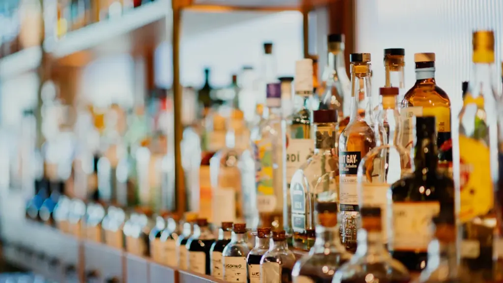 Alkoholinių gėrimų buteliai sudėti lentynose, iliustruojantys alkoholio prieinamumo temą Lietuvos visuomenėje.