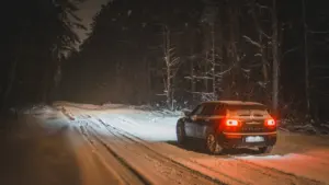 Automobilis važiuoja užpustytu miško keliu žiemos naktį, įjungti galiniai automobilio žibintai.