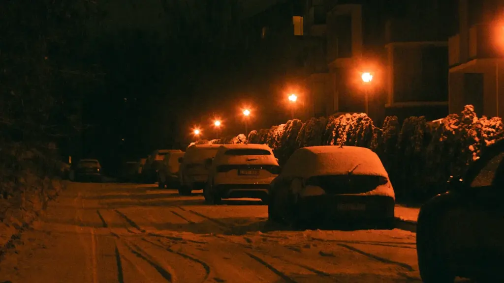 Automobiliai stovi snieguotoje Vilniaus gatvėje vėlyvą tamsų žiemos vakarą.