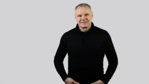Vidutinio amžiaus vyras trumpais žilais plaukais vilki juodą megztinį ir stovi prieš pilką foną.