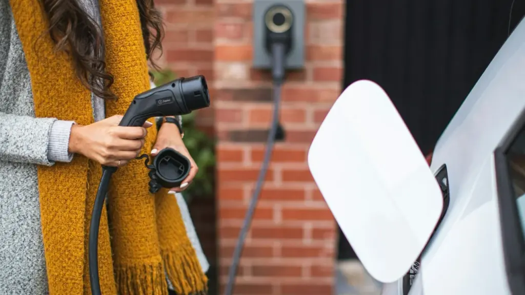 Moteris laiko rankoje elektrinio automobilio įkrovimo laidą prie baltos transporto priemonės ir įkrovimo stotelės.