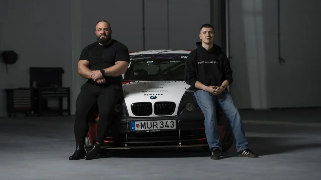 Du vyrai sėdi ant lenktyninio BMW automobilio su lietuviškais numeriais viduje, garažo aplinkoje.